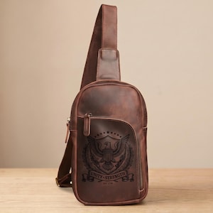 May include: A brown leather sling bag featuring a single shoulder strap and a front zippered pocket. The front panel displays a dark engraved emblem of an eagle with wings spread, a shield, stars, and the text Unity Strength Est. 1776.