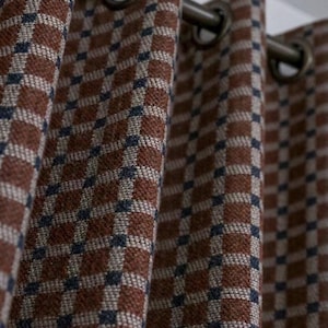 May include: A curtain panel featuring a woven checkered pattern in shades of rust brown, cream, and dark navy blue. The fabric is suspended from a horizontal rod by evenly spaced, circular metal grommets, creating deep, vertical folds in the material.