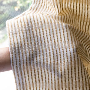 May include: A woven fabric curtain featuring a repeating vertical pattern of mustard yellow and white stripes. The textile has a textured appearance with alternating blocks of color that create a consistent linear design across the entire surface.