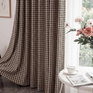 May include: A floor-length curtain featuring a brown and white checkered pattern with a grid design. The fabric hangs in vertical folds, covering a window area. The curtain is made of a woven material and displays a consistent repeating square pattern.