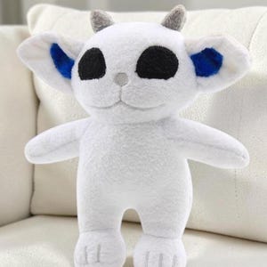 May include: A white plush toy featuring large black oval eyes, small grey horns on top of the head, and bright blue inner ears. The creature has a rounded head, pointed ears, and a small grey nose, with a soft, fuzzy texture throughout the body.