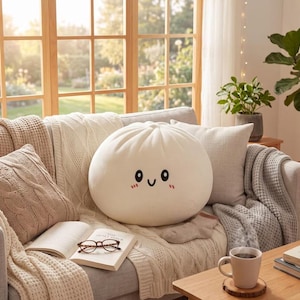 May include: A round, white plush cushion shaped like a steamed bun, featuring a simple embroidered face with black oval eyes, a curved smile, and small pink blush marks on the cheeks. The plush has a soft, smooth texture and a pleated top design.