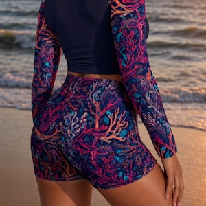 Puede incluir: Un conjunto de baño de dos piezas que incluye un top corto de manga larga y pantalones cortos a juego. El top tiene un panel trasero azul marino sólido y mangas con un patrón de arrecife de coral en tonos rosa, naranja y azul. Los pantalones cortos presentan el mismo estampado.