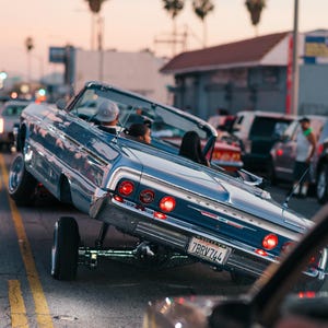 以下が含まれることがあります： クロムアクセントが施されたクラシックなブルーのコンバーチブルカー。片側が持ち上げられ、カスタムサスペンションが際立っています。車のナンバープレートは「7BRV744」です。夕暮れ時の街路で、背景にはヤシの木があります。