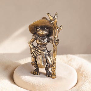 Può includere: Una piccola statuetta in metallo color ottone raffigurante una rana in posizione eretta. La rana indossa un cappello a tesa larga e una collana con un ciondolo circolare, mentre tiene in mano un alto bastone nodoso. La figura è ricca di dettagli.