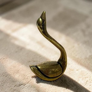 May include: A small, metallic brass-coloured figurine shaped like a swan with a long, curved neck and a smooth, polished finish. The object features a rounded body and a pointed head, capturing the elegant posture of a swan in a stationary position.