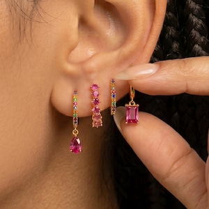 Può includere: Una collezione di orecchini dorati con pietre preziose colorate. Gli orecchini includono un paio di orecchini pendenti a goccia con una gemma rosa, un paio di orecchini a cerchio con gemme rosa e un paio di orecchini rettangolari con gemme rosa.