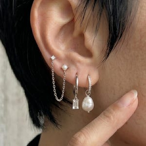 May include: A collection of silver earrings displayed on an ear. The earrings include a chain earring, a small pearl stud, a hoop earring with a rectangular gem, and a hoop earring with a pearl drop.