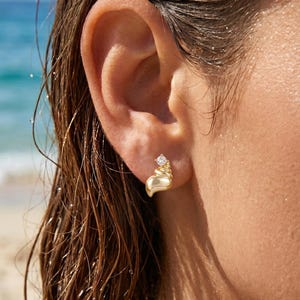 Puede incluir: Primer plano de un pendiente de oro con un diseño en forma de corazón y una pequeña gema transparente. El pendiente se lleva en una oreja, con el pelo mojado y una playa al fondo. El pendiente tiene un diseño texturizado en espiral.