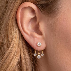 May include: A gold stud earring featuring a single round clear stone at the lobe, with three additional smaller round clear stones suspended from the base on delicate gold chains, creating a tiered drop effect against the earlobe.