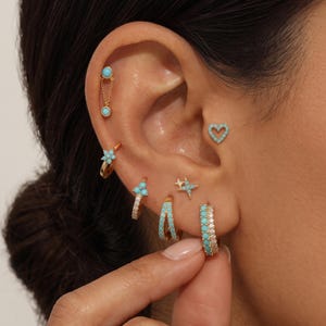 May include: A collection of gold earrings featuring turquoise stones and clear crystals. The set includes a chain cartilage stud, a heart stud, a flower hoop, a triple-stone hoop, a star stud, and a thick hoop with turquoise and crystal accents.
