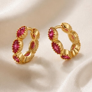 Puede incluir: Un par de pendientes de aro dorados con varias piedras de color rosa vibrante talladas en forma de marquesa a lo largo del borde exterior. Los pendientes tienen un acabado metálico pulido y un mecanismo de cierre de bisagra con diseño circular.