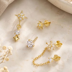 May include: A set of four gold-tone earrings featuring celestial and floral motifs with clear stones. One earring is a star-shaped drop, one is a small star stud, one is a round solitaire stud, and one is a double-post earring connected by a chain.
