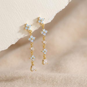 May include: Gold-toned dangle earrings featuring a floral design with light blue accents and clear teardrop-shaped pendants. The earrings have a delicate chain with small, sparkling embellishments, perfect for adding a touch of elegance.