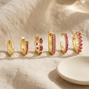 May include: A set of six gold hoop earrings arranged in a row. The collection includes one plain gold hoop, one hoop with a single row of pink stones, one with marquise-cut pink stones, one rectangular hoop with pink stones, and two circular hoops.