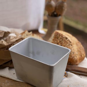 Może przedstawiać: Prostokątna, jasnoszara forma do pieczenia chleba stoi na drewnianej powierzchni, z częściowo pokrojoną bochenkiem chleba w tle. Widoczna jest również papierowa torba z mąką i mały wazon z suszonymi strąkami nasion.