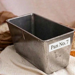 Puede incluir: Un molde rectangular de metal para pan con la inscripción "Pan No. 7" en una etiqueta blanca. El molde está sobre un paño blanco, con una bolsa de harina y una hogaza de pan al fondo. Aspecto rústico y vintage.