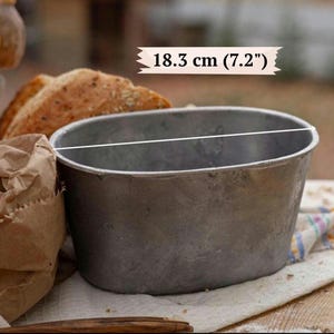 May include: An oval-shaped metal baking pan with a weathered, matte gray finish. A white line indicates a diameter of 7.2 inches, with the text 18.3 cm (7.2") displayed above the pan, showing the measurement of the opening.