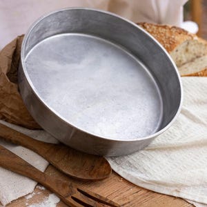 May include: A circular, silver baking tin with a broad rim, positioned on a wooden surface. Two wooden spoons and a loaf of bread are also present. The tin seems to be metal and is empty.