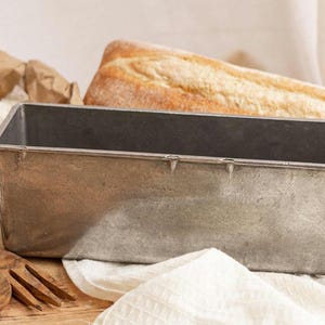 May include: A rectangular metal loaf pan, a loaf of bread, a bag of flour, and wooden utensils are arranged on a wooden surface. The loaf pan is silver and empty. The bread has a golden-brown crust. The flour is in a brown paper bag.