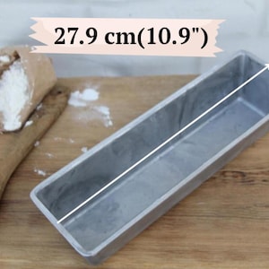 Könnte beinhalten: Eine rechteckige silberne Kastenform aus Metall mit einer Länge von 27,9 cm, zusammen mit einem Holzlöffel und einer kleinen Papiertüte mit weißem Mehl. Die Form hat eine glatte Innenfläche und eine metallische Oberfläche zum Backen.
