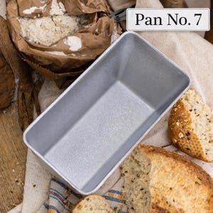 May include: A rectangular, silver-colored loaf pan with the text "Pan No. 7" in a white rectangle. The pan is empty and sits on a linen cloth next to slices of bread and a bag of flour. The scene suggests baking.