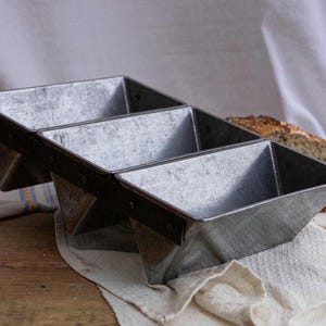 May include: A three-section metal bread pan with a rustic, industrial design. The pan is made of galvanized steel and has a dark metal trim. The angled sides and rectangular shape are ideal for baking loaves of bread. A loaf of bread is visible in the background.