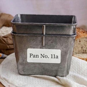 Op de afbeelding: Twee gestapelde, rechthoekige metalen bakvormen met het label "Pan No. 11a" op de voorkant. Een zak bloem en een brood zijn op de achtergrond. De pannen staan op een witte doek.