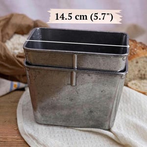 May include: A rectangular silver-toned metal bread loaf tin with a weathered finish. The tin features a vertical reinforcement rib on the side. A white graphic overlay indicates a width measurement of 14.5 cm for the top opening of the container.