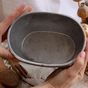 Può includere: Una teglia ovale in metallo grigio. La teglia è tenuta in mano ed è vuota. Utensili di legno e pane a fette sono visibili sullo sfondo, suggerendo un tema di panificazione.