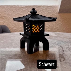 Può includere: Una lanterna nera stampata in 3D caratterizzata da un design del tetto architettonico tradizionale dell'Asia orientale con una cornice a reticolo. La struttura è sostenuta da quattro gambe arcuate e contiene una luce arancione. Il testo Schwarz è visibile.