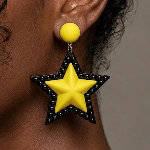 May include: A large, yellow star-shaped earring with a black border dotted with small white accents. The earring has a yellow circular stud at the top. The earring is a bold, statement piece of jewelry.