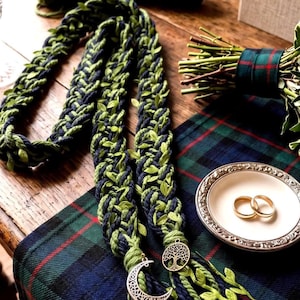 Puede incluir: Un cordón trenzado a mano hecho de hilo azul marino y verde claro entrelazado con pequeñas hojas de tela verde. Los extremos presentan dijes de metal plateado, uno con forma de media luna y el otro con forma de árbol de la vida.