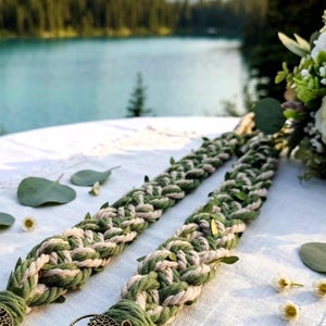 May include: Two braided cords made of intertwined sage green and cream-colored rope, featuring small green leaves tucked into the weave and a circular bronze-toned metal charm depicting a tree of life attached to the end of each cord.