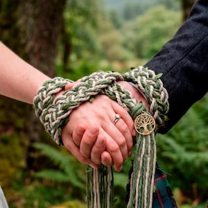 Könnte beinhalten: Ein Handfasting-Band aus geflochtenem salbeigrünen und cremefarbenen Garn, das mit kleinen grünen Blättern durchwoben ist. Das Band ist um zwei ineinandergelegte Hände gewickelt und mit einem runden, goldfarbenen Metallanhänger versehen.