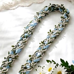 Könnte beinhalten: Geflochtene Blumengirlande mit weißen, blauen und grünen Akzenten. Die Girlande hat geflochtenes weißes Seil, blaues Band und kleine grüne Blätter. Ein Strauß weißer Gänseblümchen mit gelben Zentren und grünen Blättern befindet sich im Vordergrund.