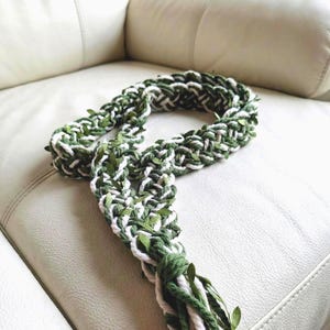 May include: A handmade, braided plant hanger in green and white, adorned with small green leaves. The macrame plant hanger is displayed on a white leather couch. The bottom of the hanger has a tassel.