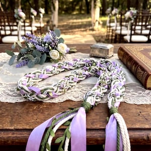 May include: A braided cord made of white rope, light purple satin ribbon, and green faux leaf vines. The cord features two long ends finished with wrapped green thread and loose ribbon tails, creating a decorative accessory with a textured, multi-layered appearance.