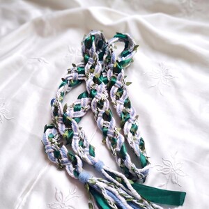 Puede incluir: Un accesorio de cordón trenzado que presenta hilos azul claro y blancos entrelazados, cinta de satén verde oscuro y pequeños detalles de hojas verdes. El cordón termina en una borla decorativa compuesta por hebras sueltas de los mismos materiales.