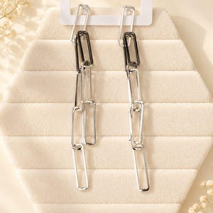 May include: A pair of dangling earrings with a chain link design. The earrings alternate between silver and black rectangular links. The earrings are displayed on a cream-colored, hexagonal jewelry display.