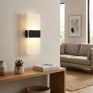 Könnte beinhalten: Moderne Wandleuchte mit rechteckigem, mattiertem Glasschirm und einem schwarzen Metallband. Die Leuchte ist an einer hellen Wand montiert und strahlt ein warmes Licht aus. Das Design ist minimalistisch und modern, geeignet für die Innenbeleuchtung.