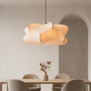 Op de afbeelding: Een grote, crèmekleurige, wolkvormige hanglamp hangt boven een eettafel. Het licht heeft een uniek, organisch ontwerp met meerdere gebogen secties. De eettafel is gemaakt van lichtgekleurd hout en er staat een vaas met gedroogde bloemen op de tafel.