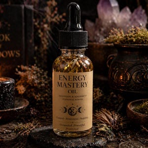 May include: A clear glass bottle of "Energy Mastery Oil" with a black dropper top, set on a dark stone. The label includes text about ascension, alignment, and personal power. The background features a lit candle, crystals, and a book.