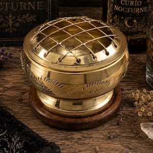 Puede incluir: Un quemador de incienso redondo de latón con una tapa de malla metálica extraíble y marcas grabadas decorativas en el lateral. El recipiente metálico descansa sobre una base circular de madera oscura y presenta un acabado pulido y brillante.