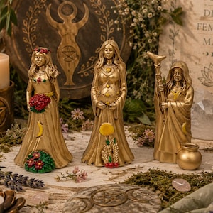 May include: Three gold-toned female figurines representing different life stages, each wearing a long gown. The left figure holds red flowers, the center figure is pregnant, and the right figure holds a staff. They are surrounded by candles and crystals.