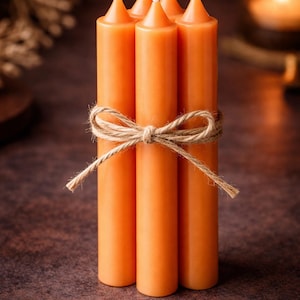 Puede incluir: Un conjunto de cinco velas de pilar naranjas atadas con hilo de bramante. Las velas tienen mechas blancas y están dispuestas verticalmente. El fondo es marrón oscuro.
