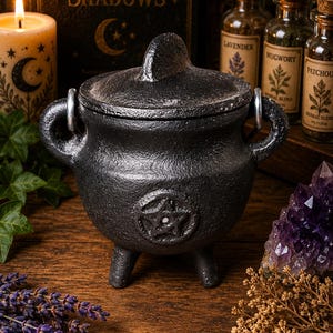 May include: A black cast iron cauldron with a matching lid, featuring a raised pentagram symbol on the front. The round vessel stands on three short legs and includes two side handles with silver-toned metal rings attached to the sides.