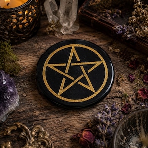 Pode incluir: Um disco preto circular com um símbolo de pentagrama dourado centrado na sua superfície. O pentagrama é composto por cinco linhas entrelaçadas que formam uma estrela dentro de uma borda circular, com um acabamento dourado texturizado.