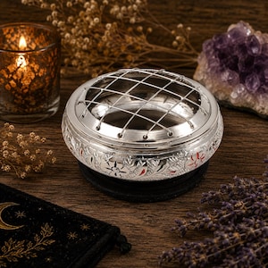 May include: A round silver-toned metal incense burner featuring an intricate floral engraving with subtle red accents and a removable wire grid top. The bowl-shaped vessel has a polished finish and sits on a dark circular base, designed for holding incense.