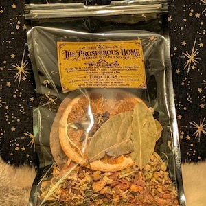 May include: A clear plastic pouch containing a simmer pot blend labeled The Prosperous Home. The contents include dried orange slices, bay leaves, cinnamon sticks, ginger root, green cardamom seeds, cloves, chamomile, calendula, and star anise.